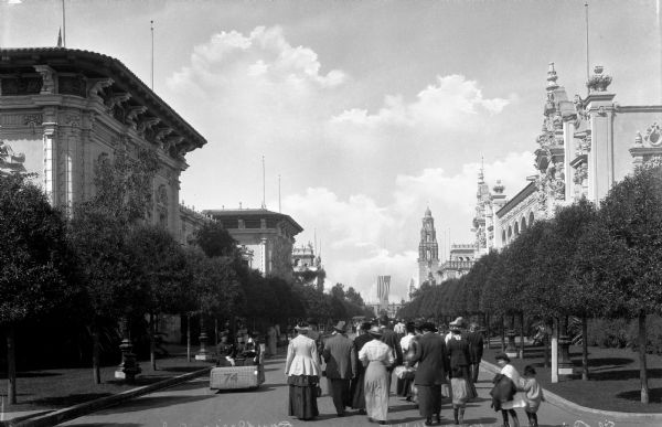 A crowd walks down a road in Balboa Park, located at the Plaza de Panama. Elaborately decorated buildings line both sides of the road.