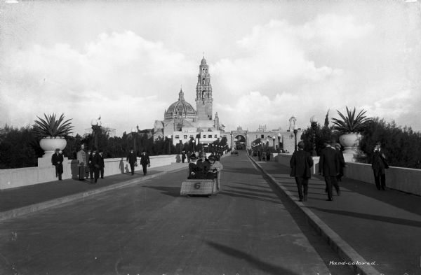 Men and women walk and drive down a street leading to the entrance of Balboa Park, established in 1868. Beyond the entrance stands the California Tower and Dome, built for the 1915 exposition.