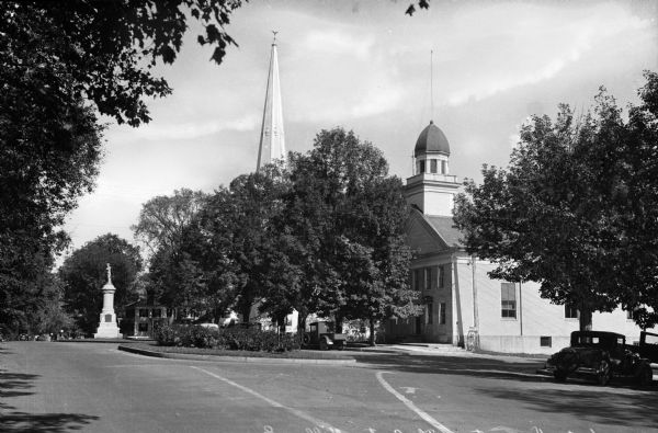 View of the center village green featuring parked cars and a monument.  A church steeple rises above trees.