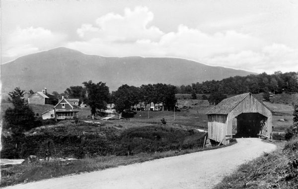 View of a covered bridge, looking toward South Lincoln. Dwellings are in the distance, and mountains are in the background.