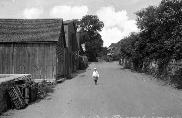 A boy walks down Shore Road. Wooden sheds and a country home stand along the roadside.