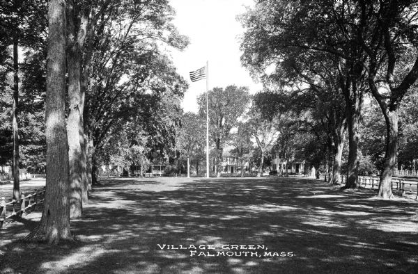 View of the village green featuring rows of trees and a flagpole surrounded by a fence.