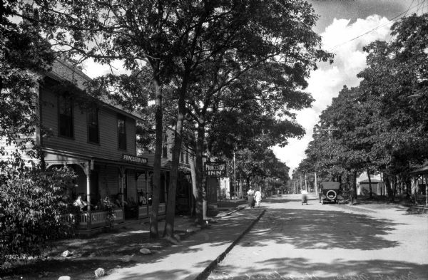 View down tree-shaded Highland Avenue featuring the Princeston Inn, a boarding residence.  Men sit on the porch as other men and women walk down the sidewalk past a parked automobile.