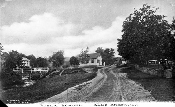 View of a small public school house in a country setting.  A dirt road leads past a stream and a wooden bridge toward the school house.