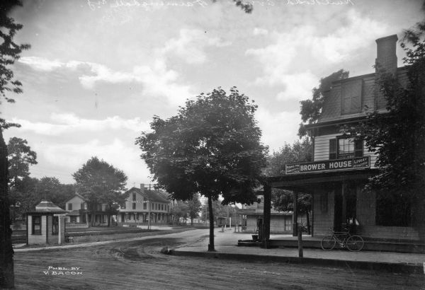View down Railroad Avenue, a curving country road lined with inns.  The Brower House, established by Charles Chauncey Brower, is visible in the foreground at right.  On the left stands American Hotel, built in 1868. Published by V. Bacon.