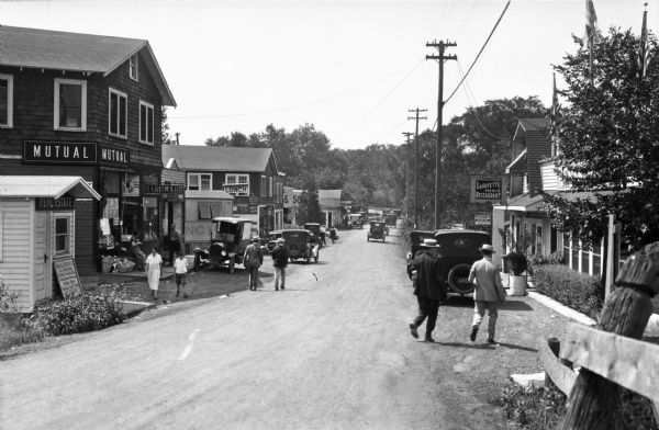 Pedestrians pass parked cars along a narrow dirt road lined with shops.