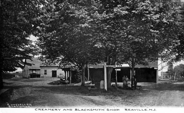 Six children and a woman pose under a tree near a creamery and blacksmith shop. A sign to their left reads: "Flemington 4 M" and "3 Bridges 3 M." Published by E. Vosseller Stationer.