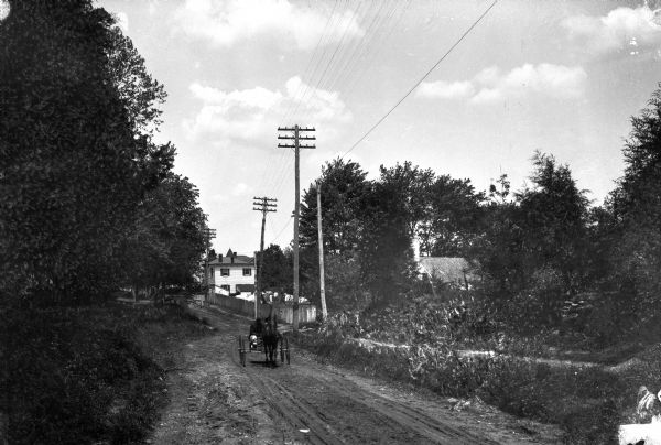 A man steers a horse-drawn carriage down a sunken dirt road in a rural setting.  Dwellings stand at right.