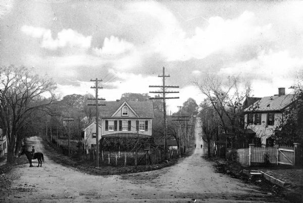 View of an intersection of two dirt roads, East Market Street and Lafayette Street.  A man, horseback, can be seen on the left, while two people walk past dwellings down the road at right.