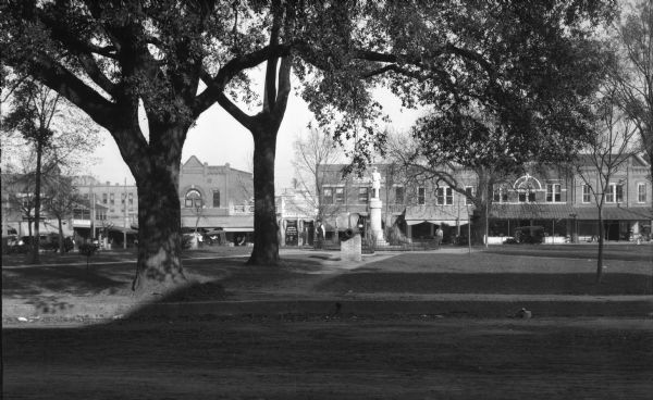 View of Fort Hughes featuring a park or town square decorated with a cannon and a statue. A street lined with storefronts is visible across the lawn.