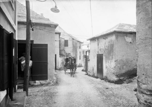 A man steers a horse-drawn carriage down an alley as two others look on from a doorway. The photograph was taken in Saint George, Bermuda.