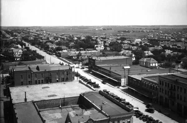 Elevated view of the town and surrounding area.  The brick building at the photograph's bottom center is labeled "Geren, Rish Art Studio" and was built in 1898.  A necessity store, dry goods company, and a furniture company can be seen across the street near a line of parked automobiles.