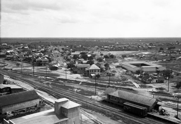 Elevated view of town buildings with railroad tracks running through the town. A railroad depot stands in the foreground.