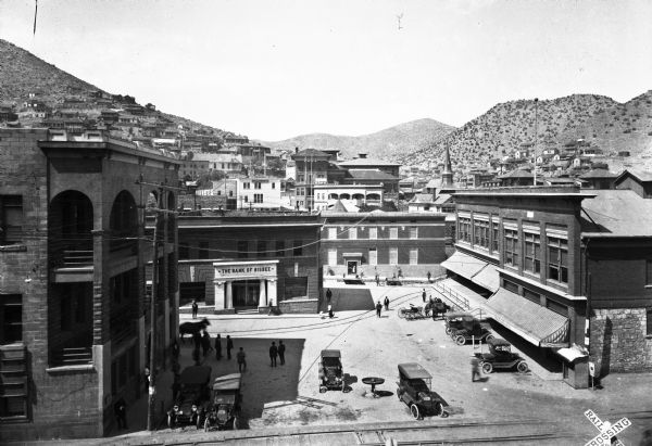 Elevated view of streets, stores, the Bank of Bisbee, and dwellings built into the hillside. Pedestrians, automobiles, and horse-drawn vehicles are visible in the foreground.