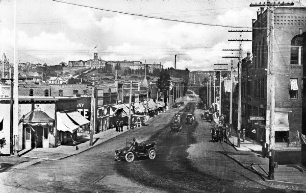 View of Main Street, featuring shops, cars, and carriages.  Washington State College, founded in 1890, can be seen on a hilltop beyond the street.