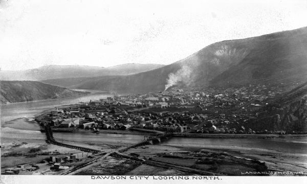 General view looking north of Dawson,Yukon, Canada. Features a river, bridges, and the city's buildings.  Published by Landahl's Emporium.