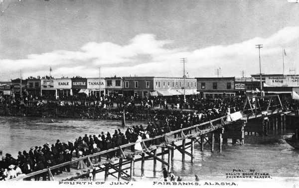 A crowd gathers on the pier and on the shore during a Fourth of July festival.  Shops can be seen beyond the wharf.  Published by Hall's Bookstore.