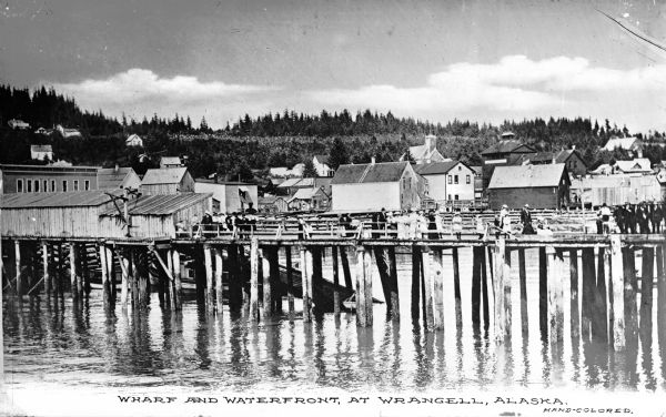 Men and women stand on the pier at the wharf and waterfront. Buildings are visible in the distance.