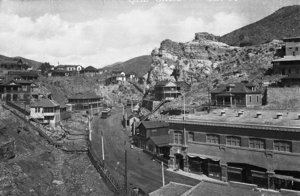 View featuring stores and dwellings built into hillsides. Castle Rock  appears on the right side of a road.  Opposite the landform, the Castle Rock Lodging House can be seen; it was built in 1895.