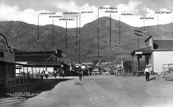 View down Tennessee Avenue, with the locations of mines marked in the hills in the distance. Men walk past shops on either side of the avenue.