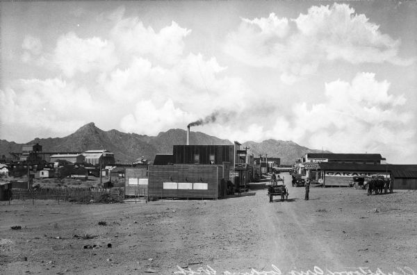 View down a dirt road featuring a repair shop and other shop buildings.  Men travel past the buildings by horse and carriage and by automobile.