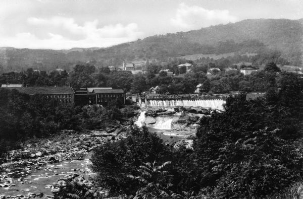 View of a landscape with a rocky stream in foreground and town set in countryside in the distance. Shelburn Falls can be seen among industrial and residential buildings.