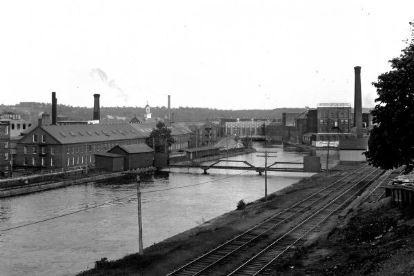 View of the First Level Canal lined with industrial buildings featuring a paper mill,founded in 1882, on the right. A sign on the buildings reads, "Whiting Paper Company, Ledgers, Linens, Bonds, and Fine Writing Papers." Railroad tracks can be seen on the right bank of the canal.