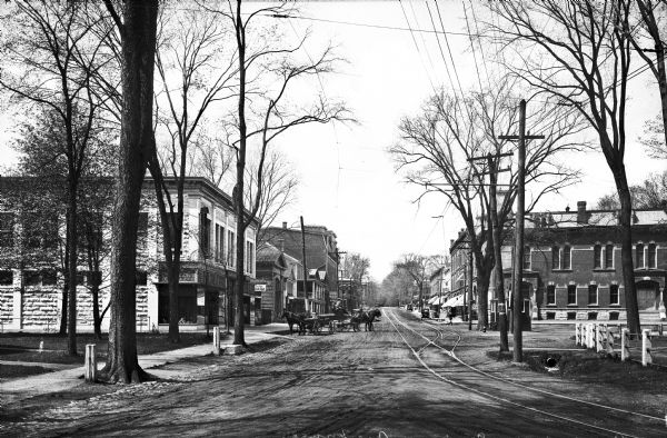View down Lower Main Street featuring shops, a horse and carriage, automobiles, and trolley tracks.