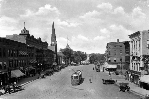 View down Main Street from City Hall featuring horses and wagons, automobiles, and street cars.  Stores line either side of the street.