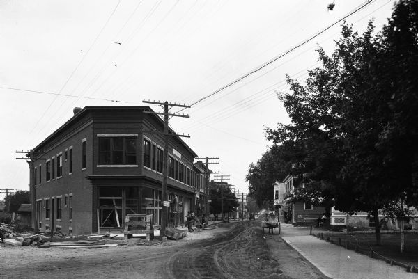 View down Prospect Street, a dirt road, featuring the construction of a brick corner building.  Two men look on from the steps of their home across the street.