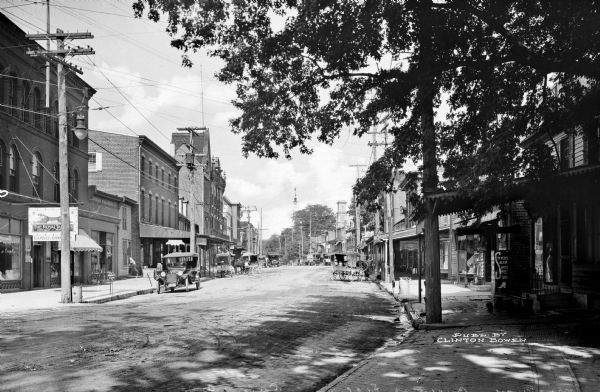 View down Broadway featuring carriages and automobiles parked near storefronts.  Published by Clinton Bowen.