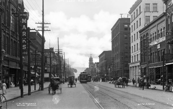 View down State Street featuring cable cars, horse-drawn carriages, and bicycles.  Pedestrians pass by shops and the Marine National Bank on the right, known as the 'marble front.'  Published by J.M. Stearn's Book Store.
