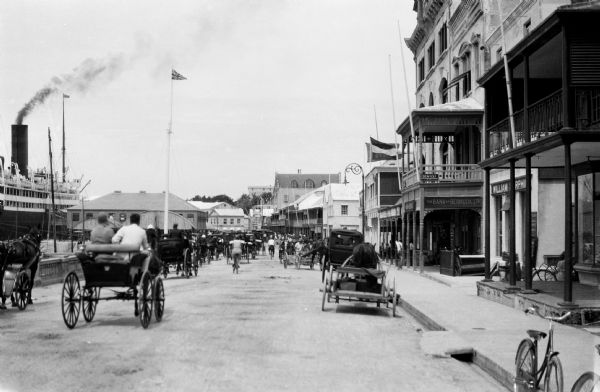 View down a busy Bermuda street featuring shops and horse-drawn carriages.  A large ship is docked at left.