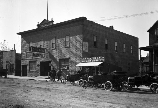 Street scene in front of a garage labeled Dolloff Block, 1906. Men stand outside Moore Motor Company and J.M. Deeter & Son's Pop Corn Crispetts stand. A sign on the side of the garage reads, "The Next Car Leaves in Ten Minutes for the Old Settlers Picnic."