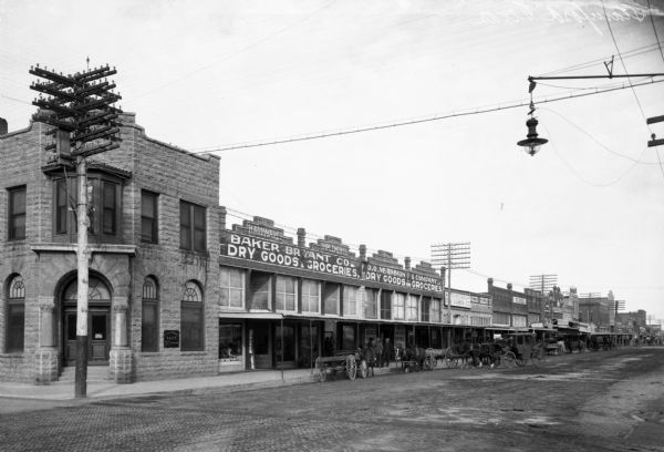 View down a town street featuring dry good and grocery stores including Baker Bryant Co. and D.O. McRimmon & Co.  Horse-drawn carriages park along the street.