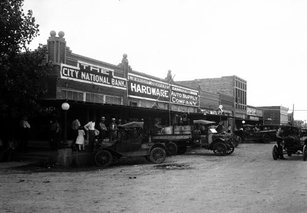 Automobiles line unpaved Main Street which features the City National Bank, L.B. Wright Hardware Company, and Carrol Bros. Auto Supply Company.