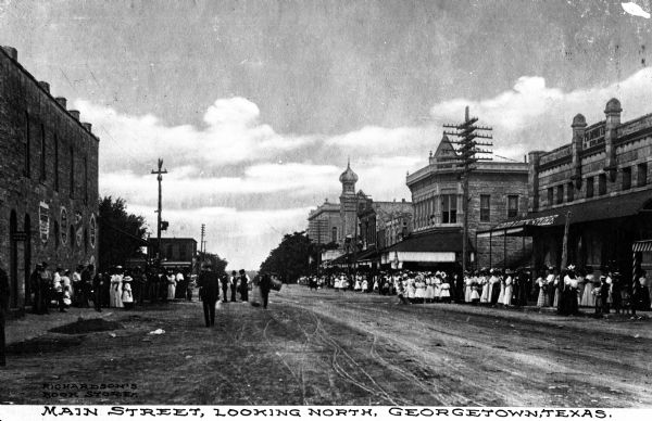 View down Main Street, looking north.  Groups of people congregate near storefronts.  The stone building at right was constructed in 1891.