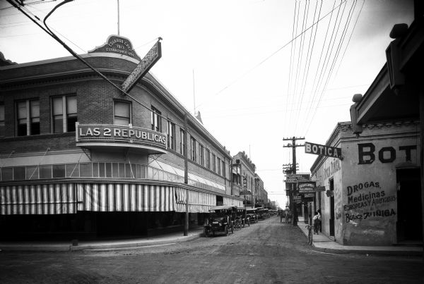 View down a street featuring stores and small businesses with signs written in Spanish. On the left corner, Eduardo Cruz' department store can be seen, built in 1913 and established in 1894.  A drugstore stands across the street. A botica, pharmacy and drugstore is on the right.