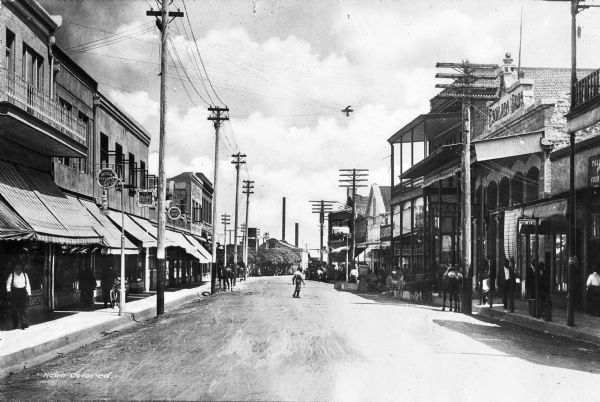 View down Morley Avenue, lined with stores and small businesses.  A stone building at right is labeled 'Escalada Bros.'; it was established in 1892 by the Spaniard Manuel Escalada.