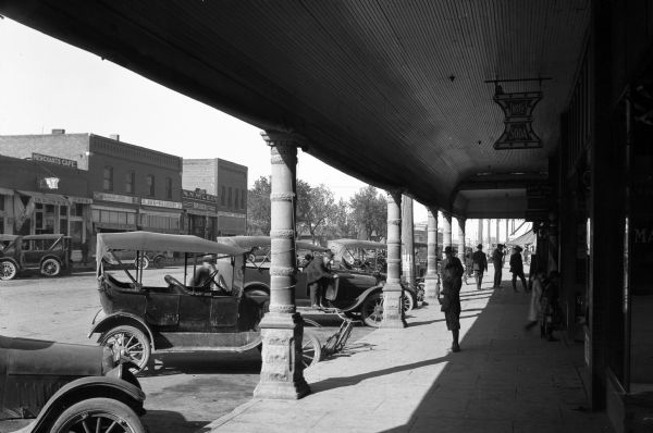 View down a city street from a colonnaded walkway.  Automobiles park outside a store advertising drugs and soda, Wade Bros. Land Company, and Henry C. Rogers Attorney Office.