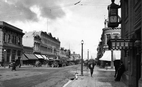 View down a city street.  Men and women walk past stores, restaurants, and Monterey County Bank, established in 1890.