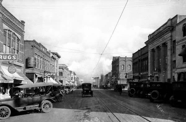 View down Main Street. An automobile drives past stores, cafes, and First National Bank; the bank was organized in 1905 and erected in 1911.  In the background, a banner advertises a Poultry Show, taking place January 6 - 10 on the corner of 6th and Central.