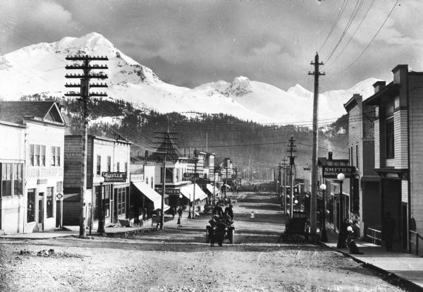 View of First Avenue, the main street in town. Stores, restaurants, and lodging houses stand on either side of the street. Snow-covered mountains rise in the background.