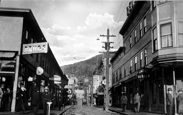 View down Front Street in the business district. Hotel Revilla, which burned in 1924, stands at the right. Men walk past stores on the left.