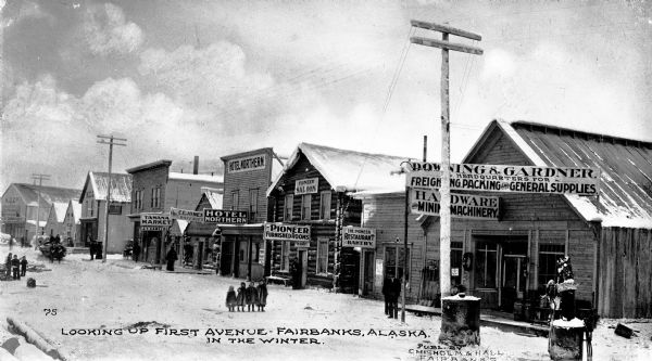 View down business-lined First Avenue. Pedestrians are walking along the street. Caption reads: "Looking Up First Avenue — Fairbanks, Alaska. In The Winter." and "Publ. By Chisholm & Hall Fairbanks".