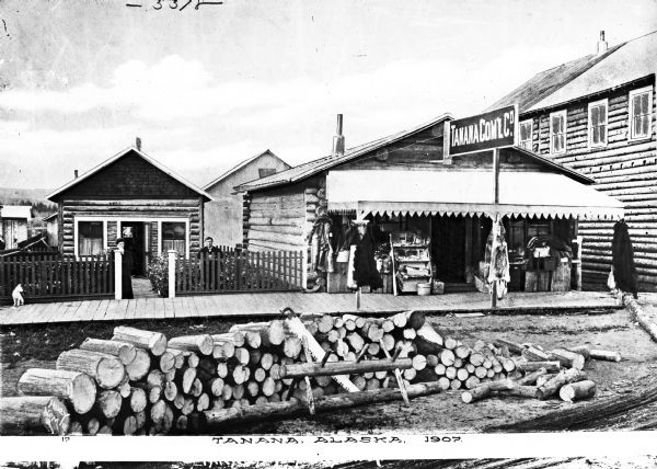 A couple stands outside their dwelling adjacent to Tanana Commercial Company, established in 1880.  Stacks of lumber and a large saw are visible in the foreground.