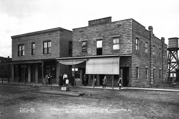 View of two buildings that face Courthouse Square. Men, women, and children stand outside the Baldwin Drug Company at right. Published by Baldwin Drug Co.