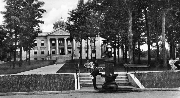 View of the Sullivan County Courthouse, built in 1909. Three young boys read a newspaper on a park bench in front of the building while a man drinks from a nearby fountain.