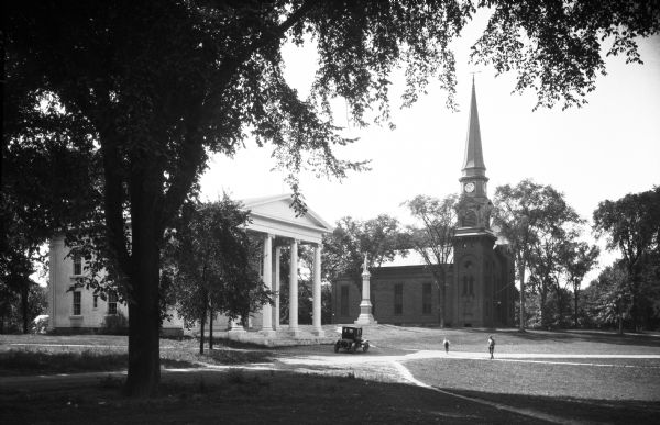 View of Branford Town Hall and First Congregational Church, two Greek Revival buildings are located near a monument.  The town hall was built in 1857 and features a colonnaded porch.  Beyond the statue stands the church building with its clock tower, constructed in 1843.