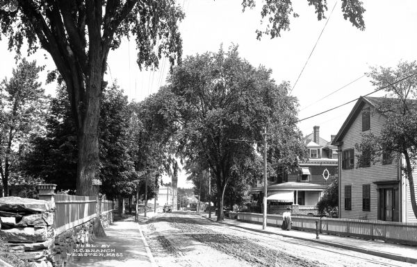 View down a residential street lined with picket fences.  An automobile and trolley car travel down the road.  Published by H.C. Branch, Webster, Massachusetts.
